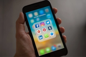 Hand holding a smartphone displaying a folder of social media apps like Facebook, Instagram, and LinkedIn—symbolizing strategic SMM campaigns that connect brands with local audiences-ABIQA